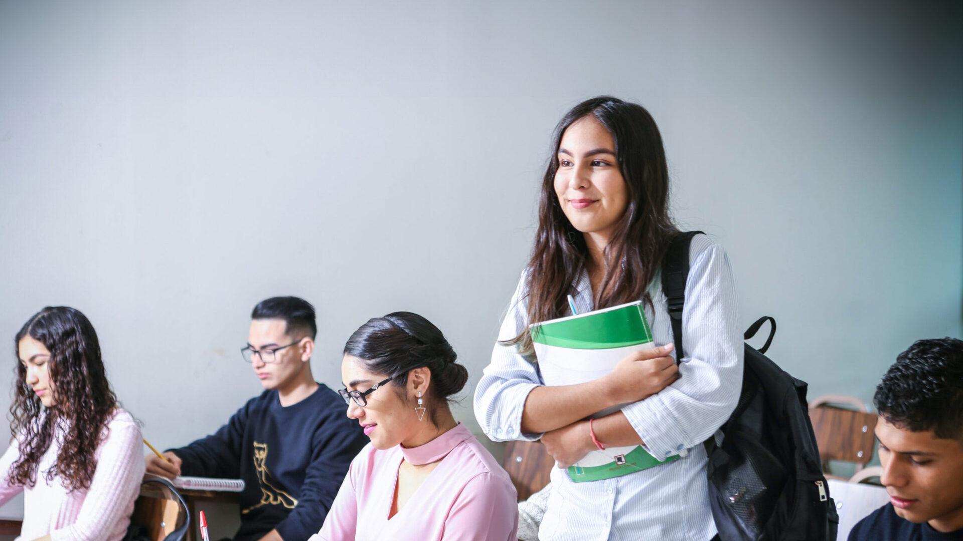 A student clutches her textbooks and stands among her seated classmates.