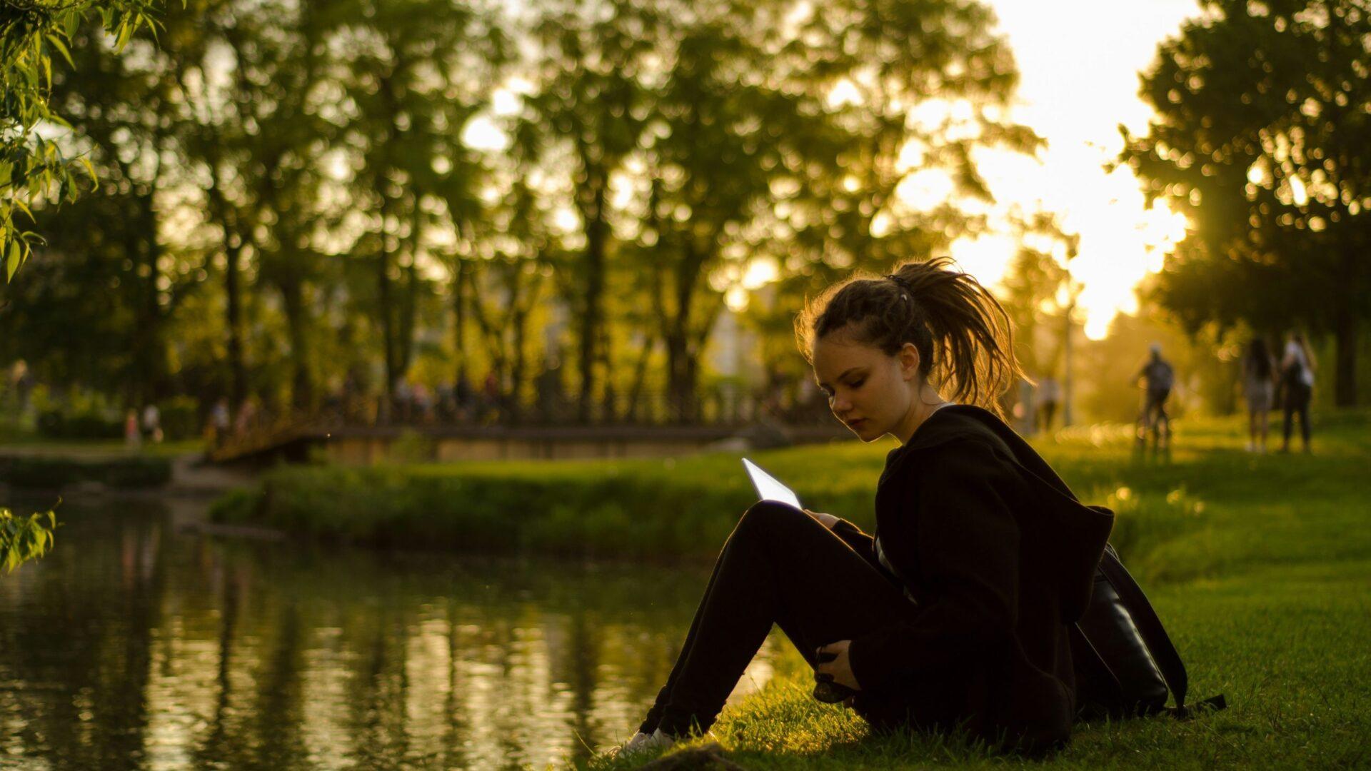 A young woman sitting in the grass near a lake at sunset.