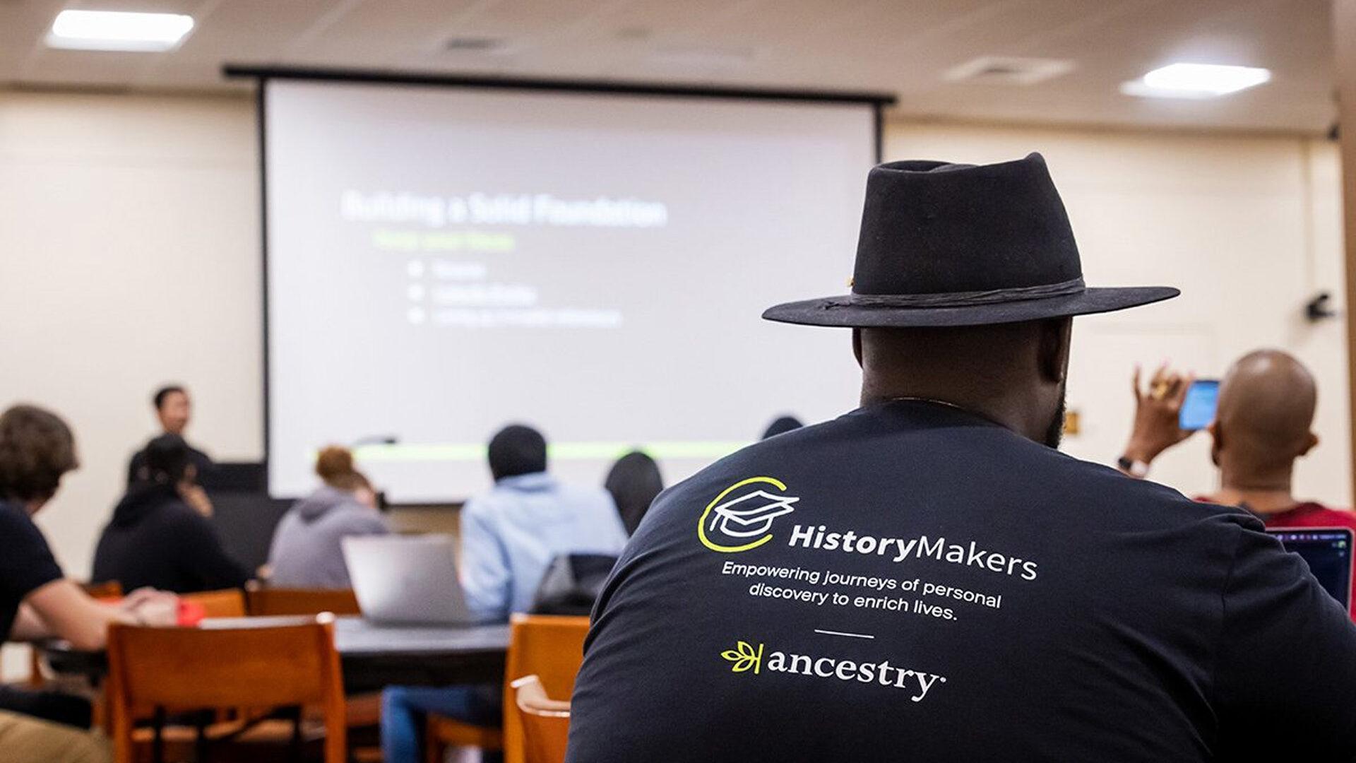 A man wearing a hat and a shirt reading "HistoryMakers," sitting in a classroom.