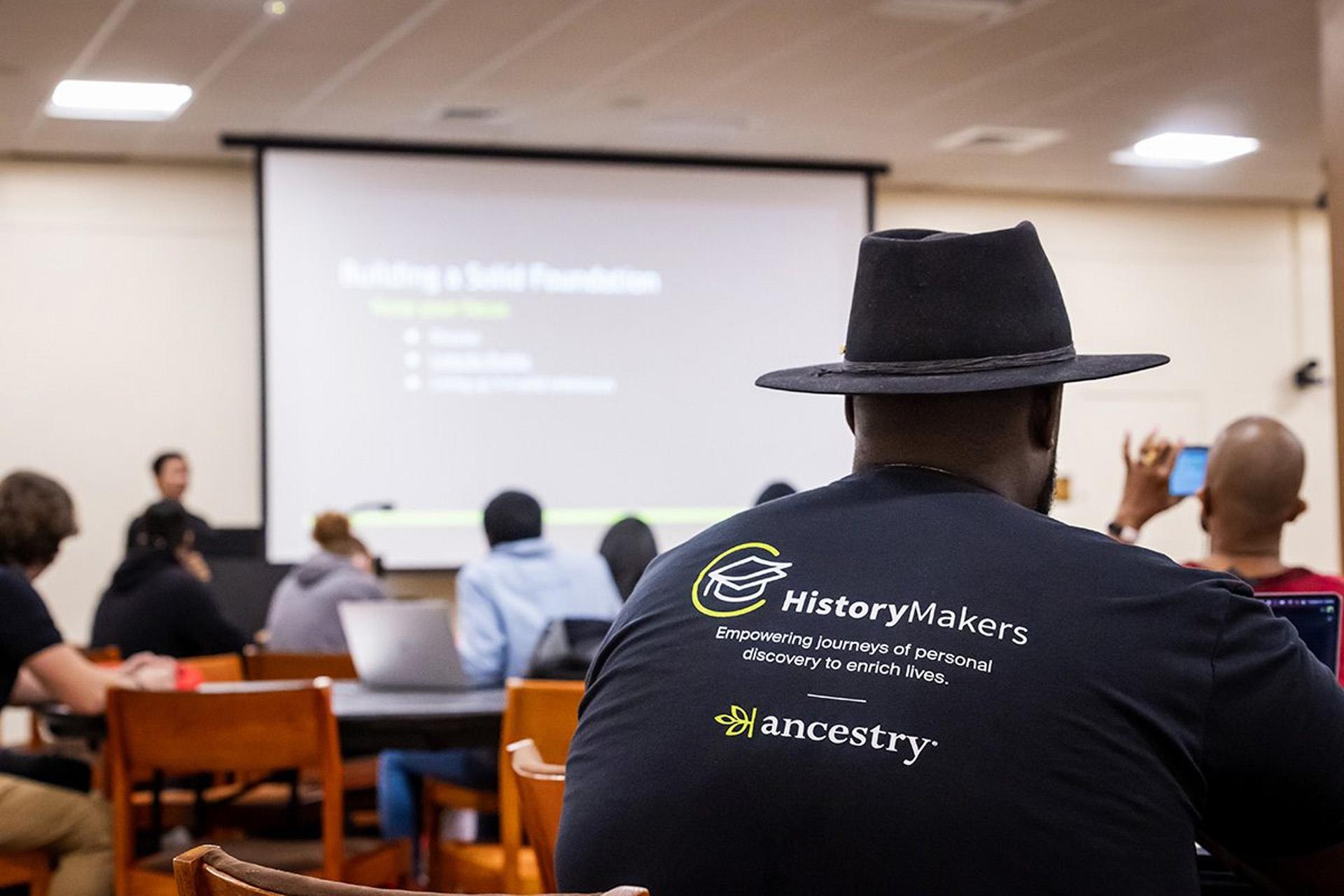 A man wearing a hat and a shirt reading "HistoryMakers," sitting in a classroom.