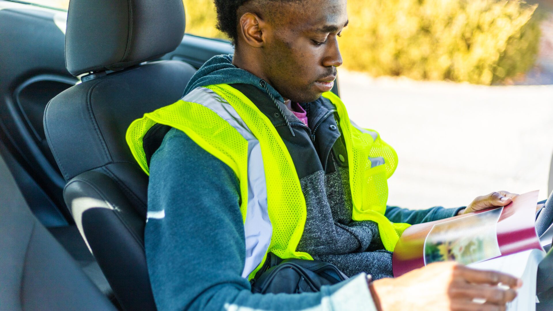 A young man wearing a high-visibility jacket studies in his car during a break from work.