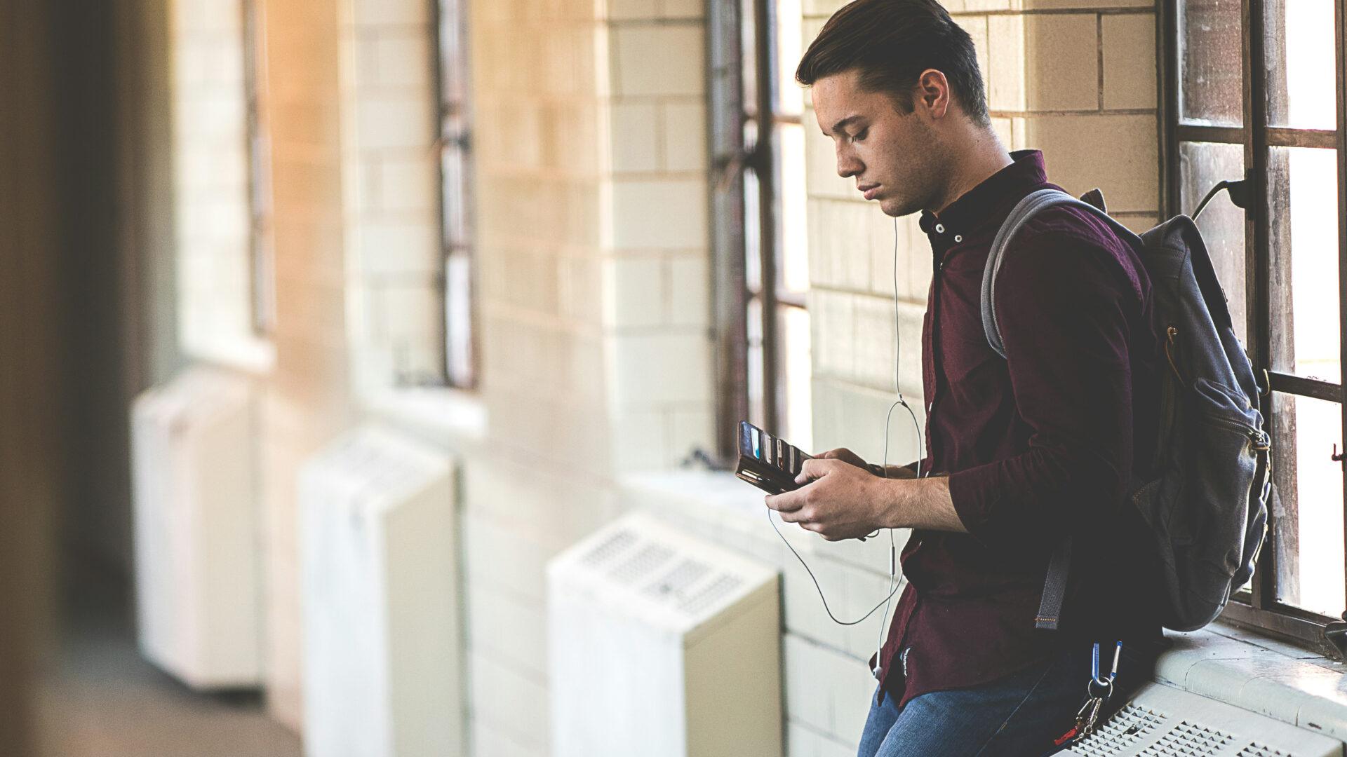 A student leaning against the wall in a hallway listening to music on his phone.