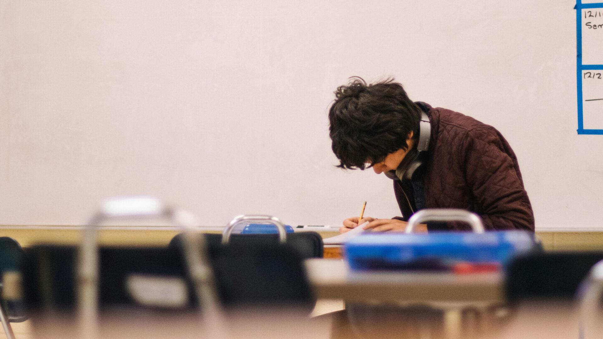 A student hunches over his books, studying in front of a whiteboard.