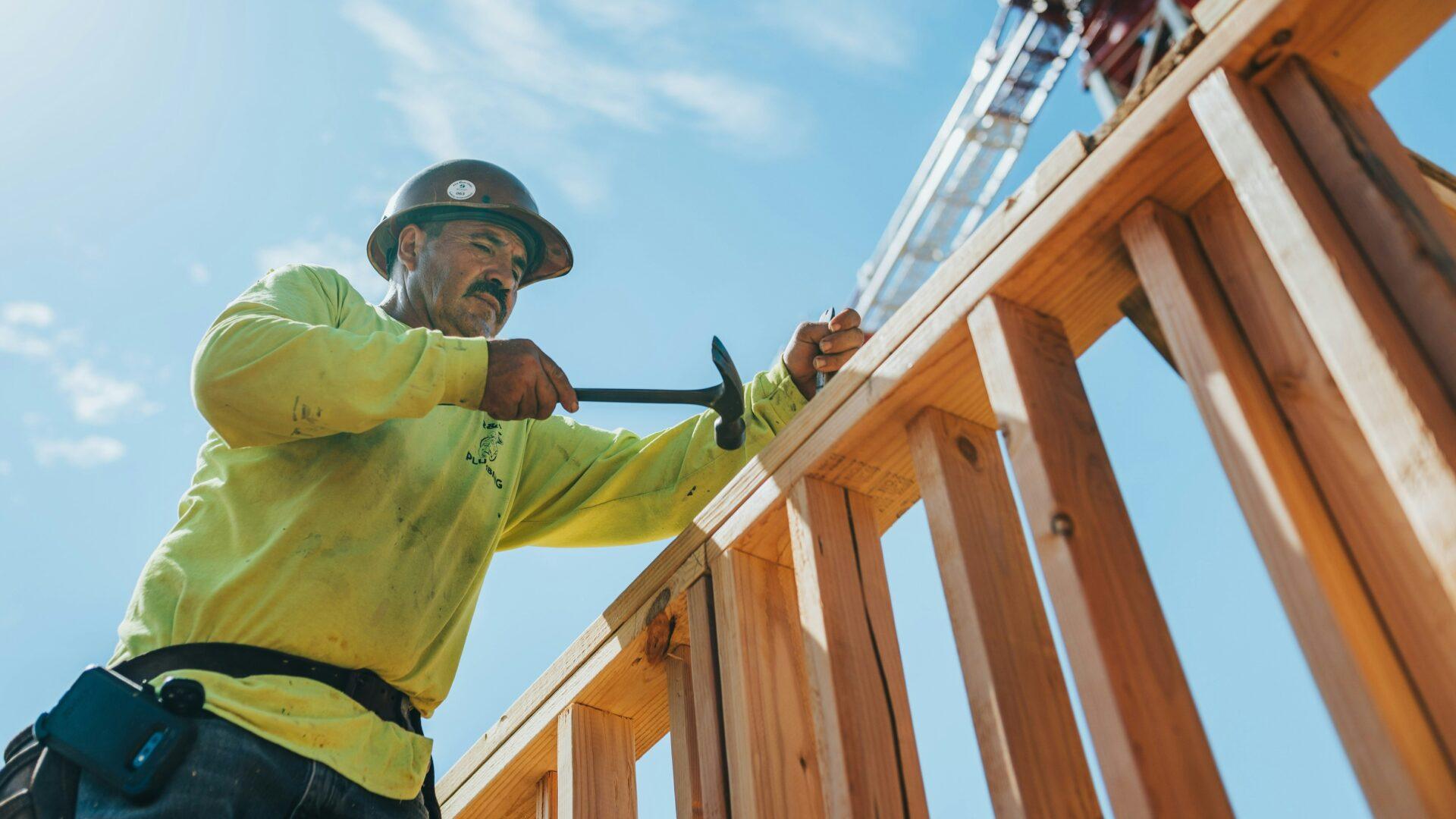 Construction worker in a high-vis shirt working on a wooden frame with a hammer outside in front of a bright blue sky