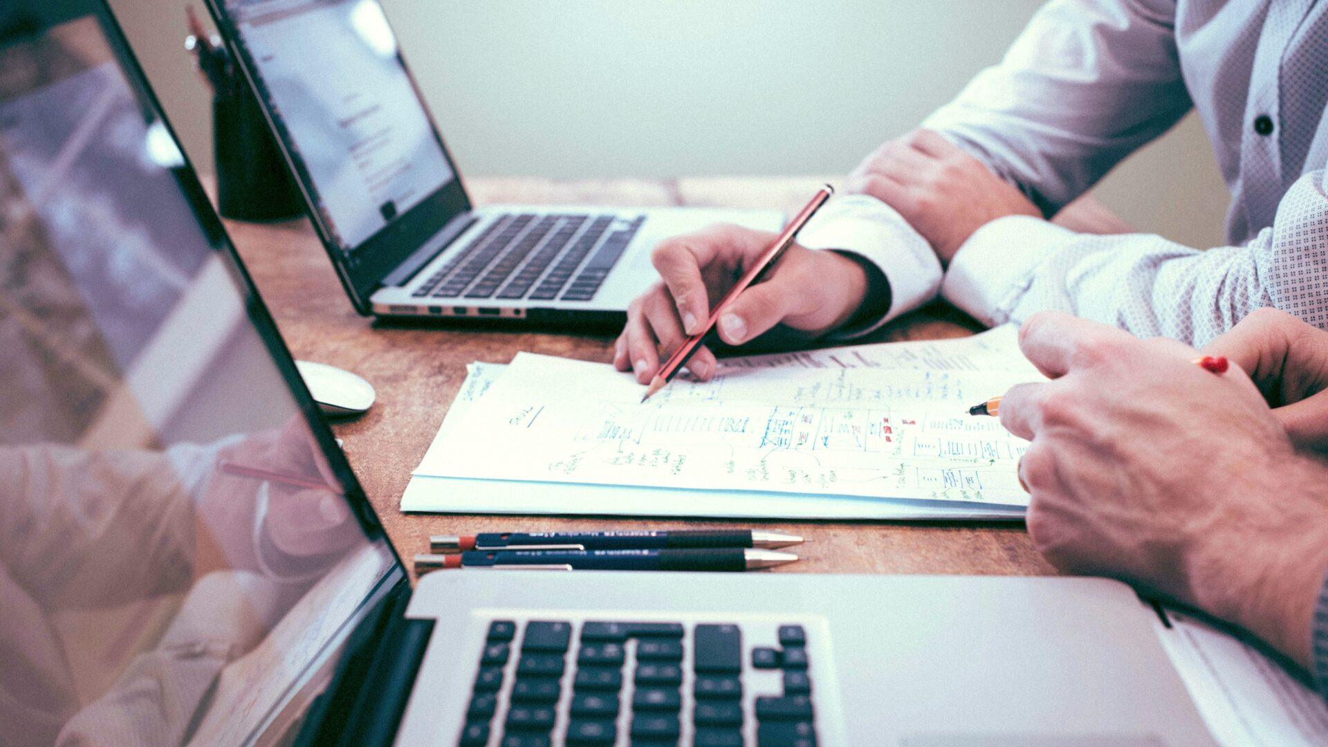 Two people working on laptops in an office, one of them holding a pencil and writing notes on a document