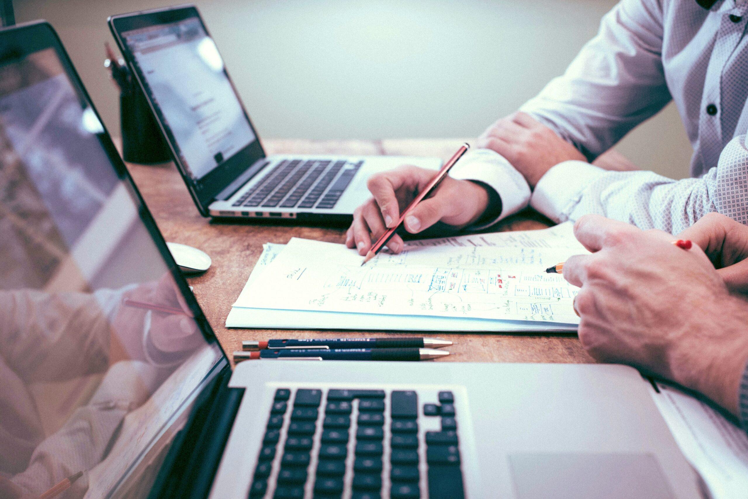 Two people working on laptops in an office, one of them holding a pencil and writing notes on a document