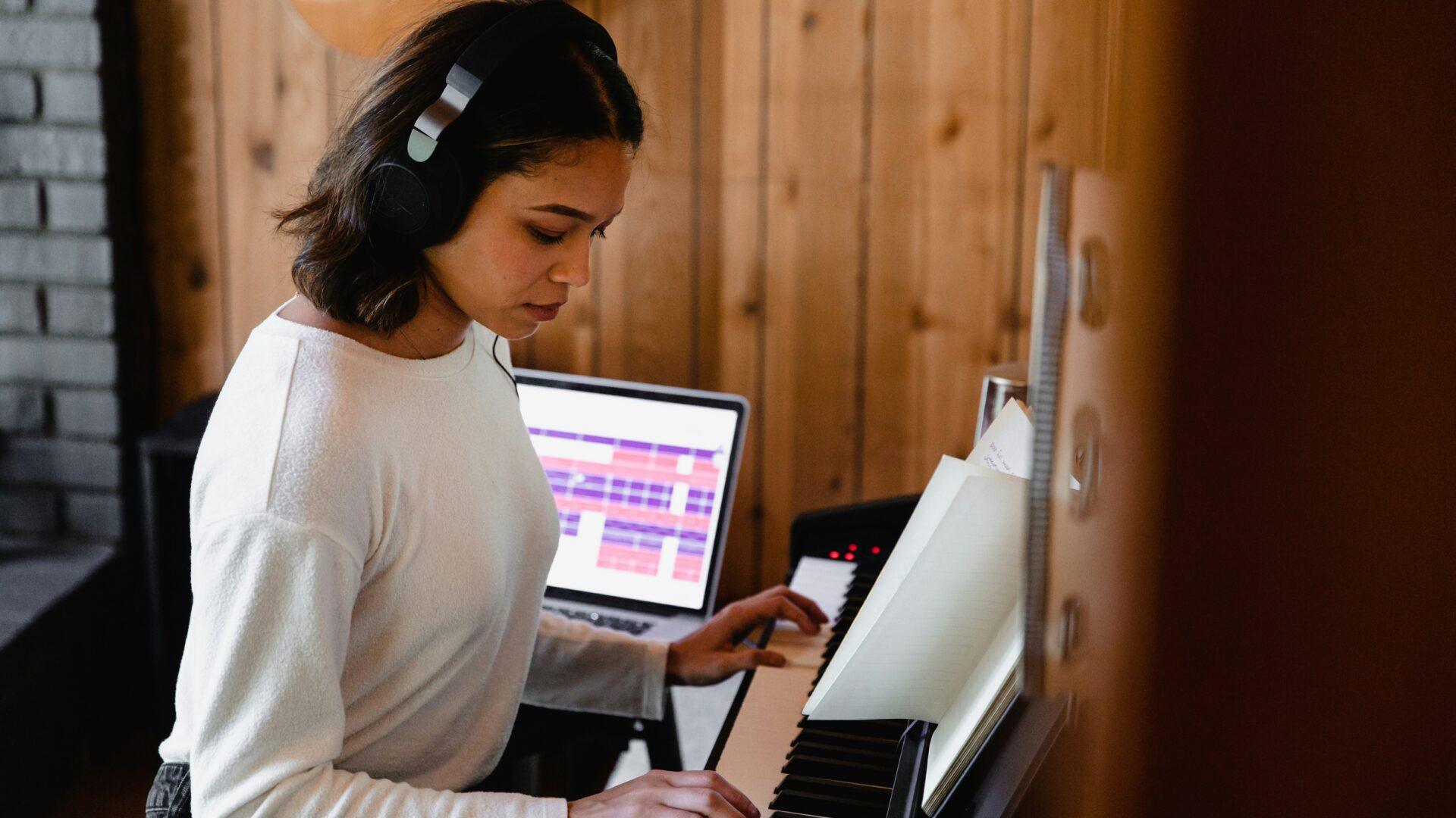 Young female music student wearing over-the-ear headphones, playing music on an electric keyboard. A computer in the background has music editing software on the screen.