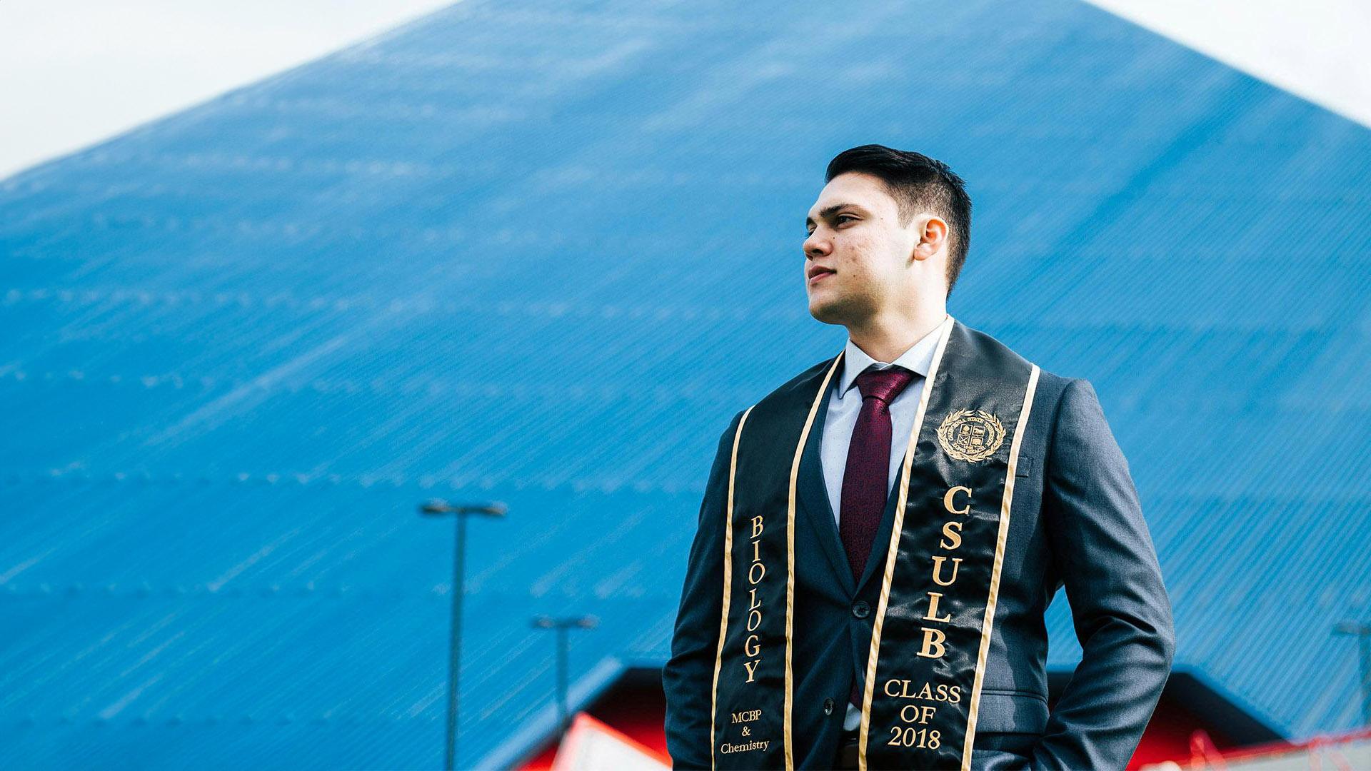 A graduate with a sash stands in front of a glass pyramid.