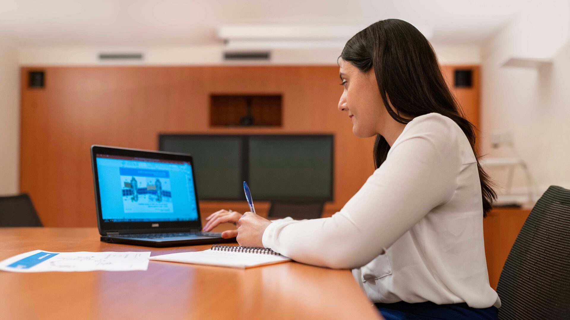 A woman sitting at a conference table reading from her laptop and taking notes.