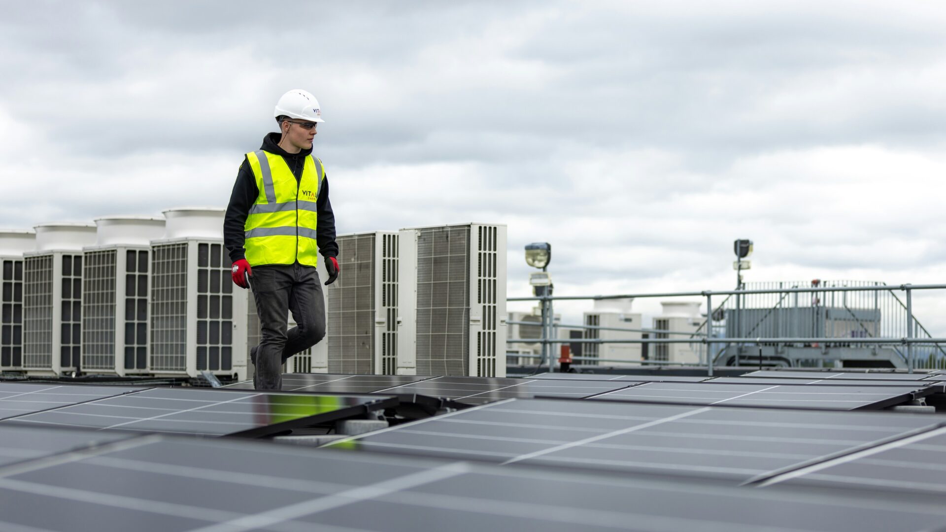 A you man in a hard hat and high-vis vest surveys a rooftop full of solar panels.