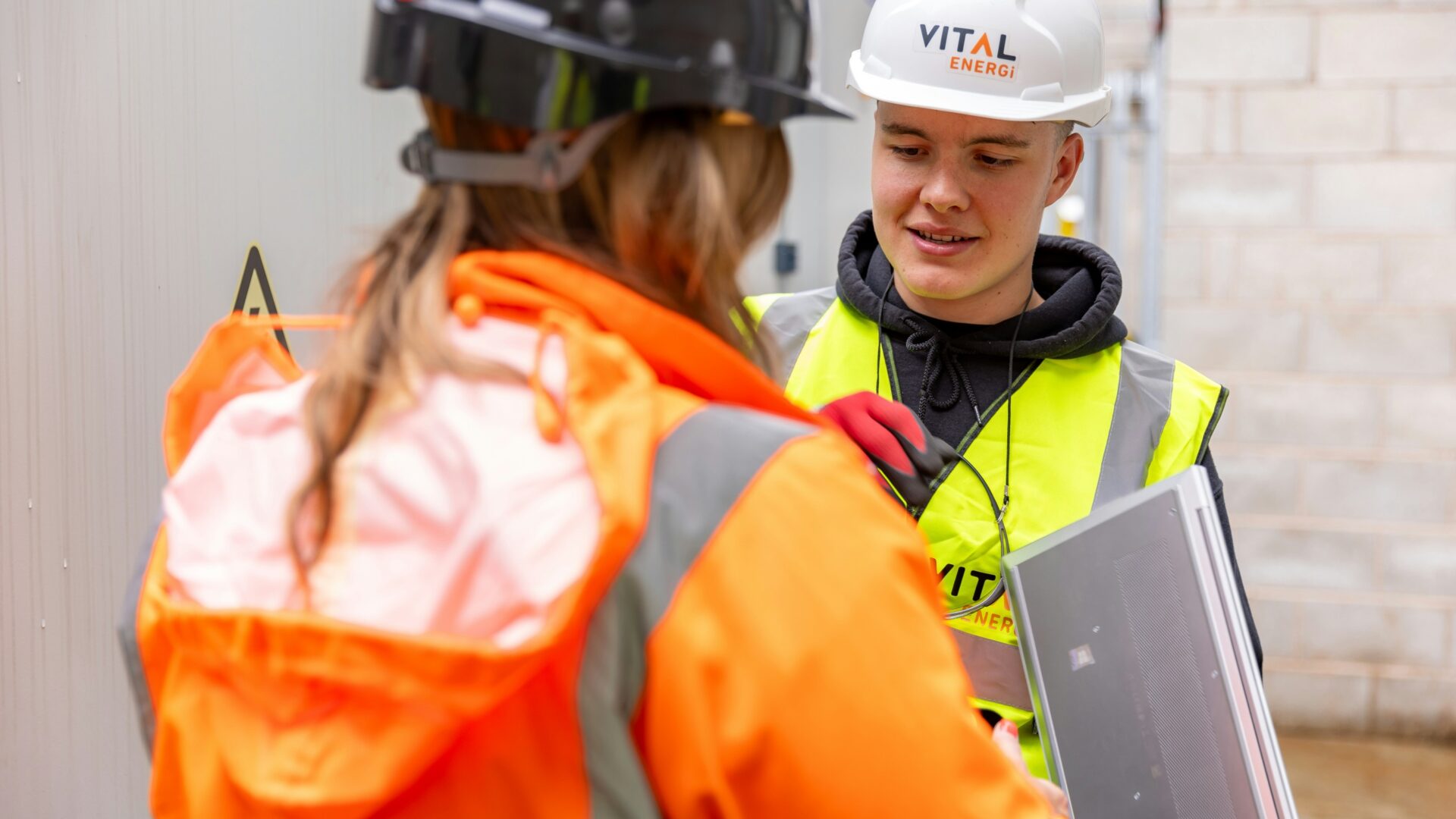 A young man and woman both wearing high-vis vests and hard hats discussing work.