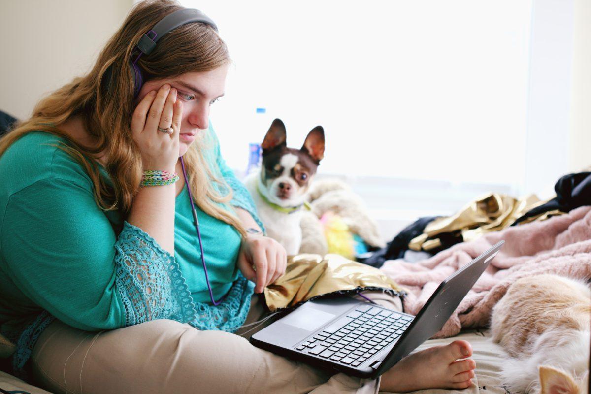 young woman sits on her bed with her dog looking at her laptop