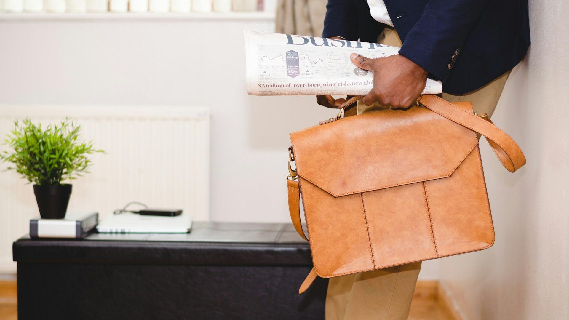 man holding brown leather bag and business section of newspaper