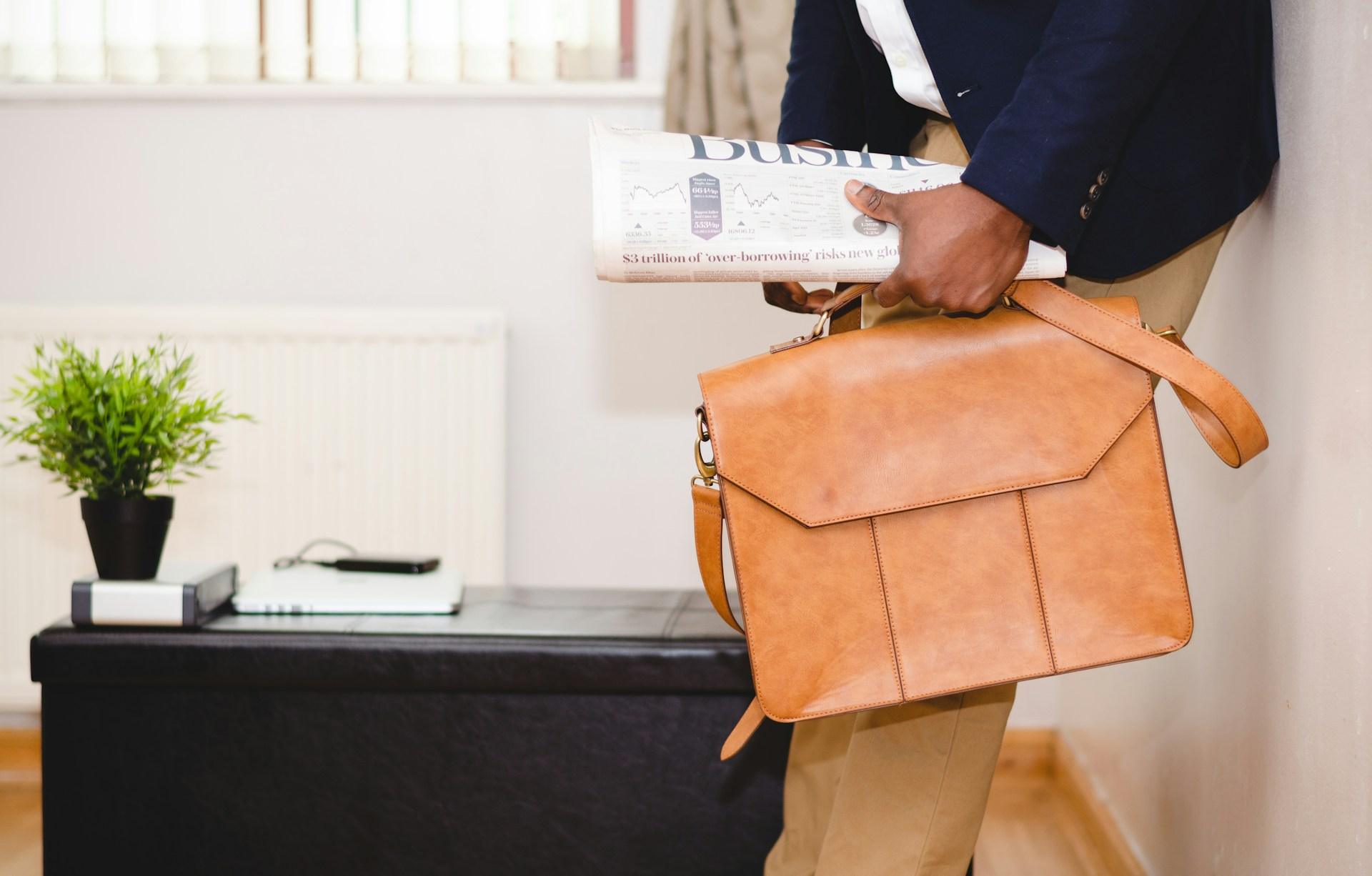 man holding brown leather bag and business section of newspaper