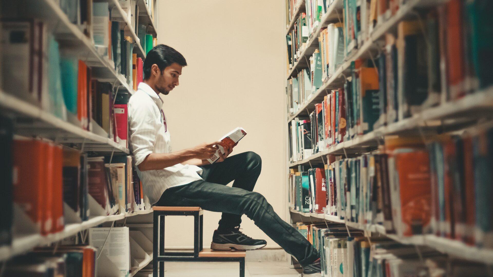 a male student leans back in a library and reads a book from the shelf.