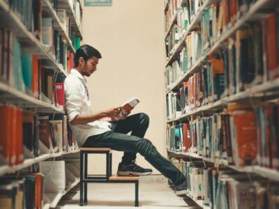 a male student leans back in a library and reads a book from the shelf.