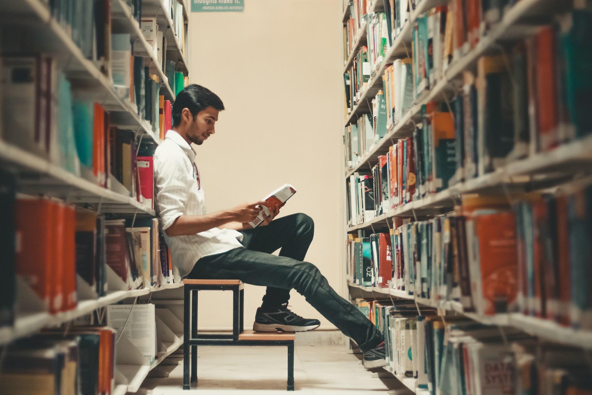 a male student leans back in a library and reads a book from the shelf.