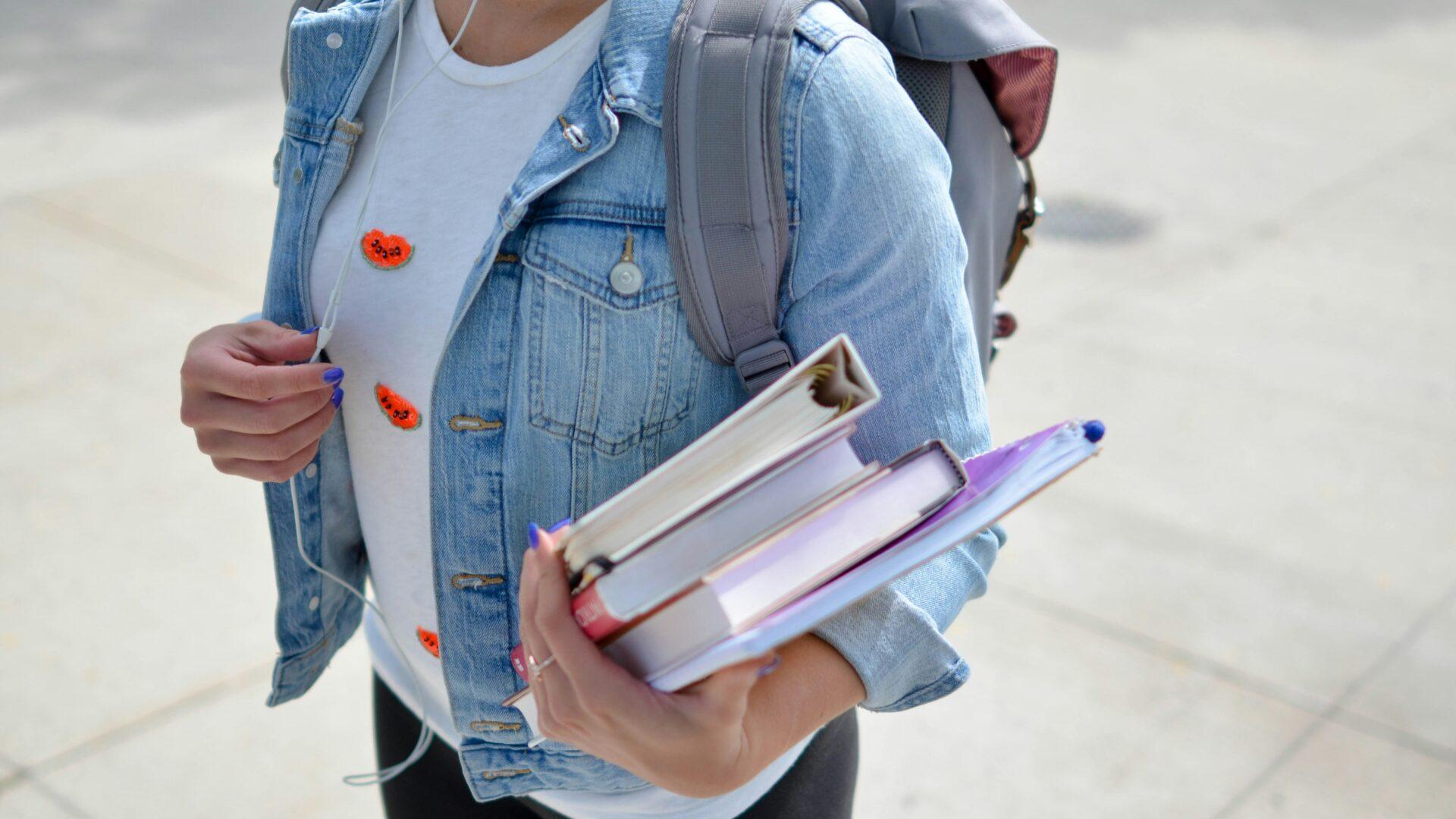 woman wearing a blue denim jacket and gray backpack and carrying school books