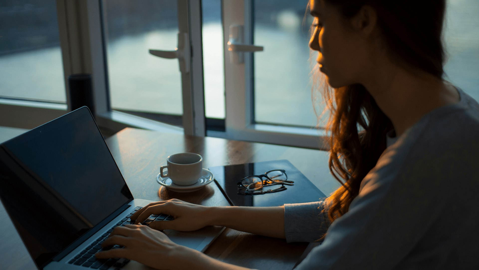 woman in low light sits at a laptop with a cup of coffee