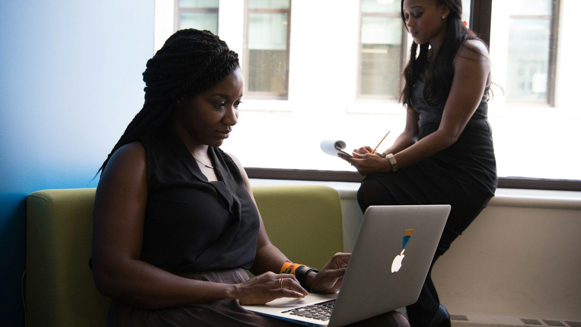 Two black women in an office, the one in front typing on a Macbook