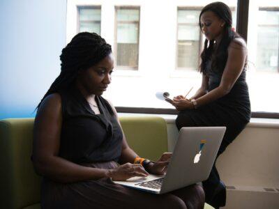 Two black women in an office, the one in front typing on a Macbook