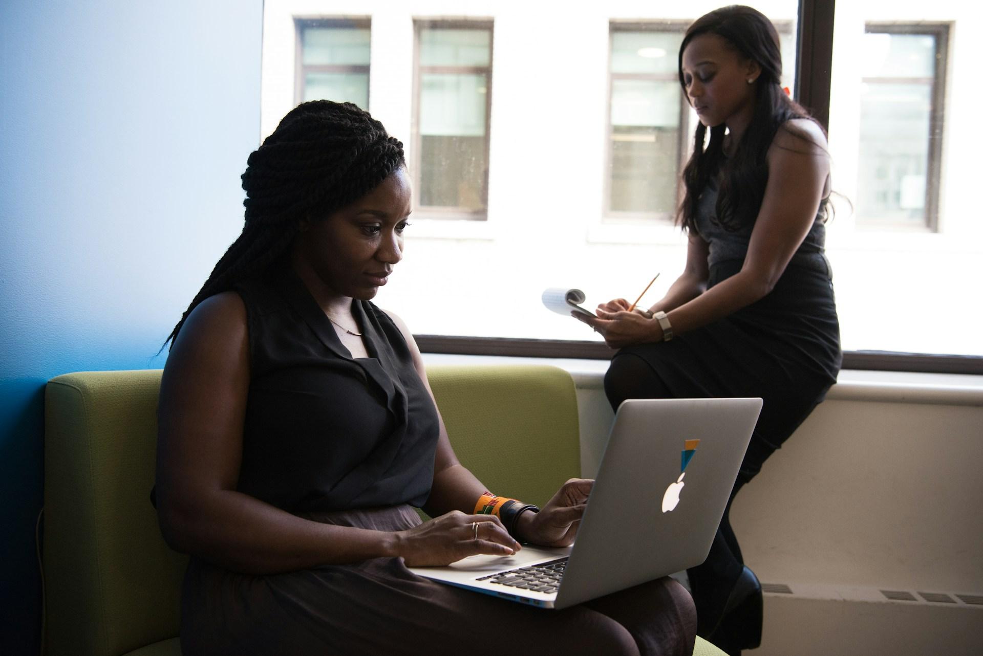 Two black women in an office, the one in front typing on a Macbook