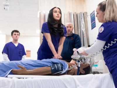 Female nursing student giving chest compressions during a CPR lesson in a lab classroom on a college campus.