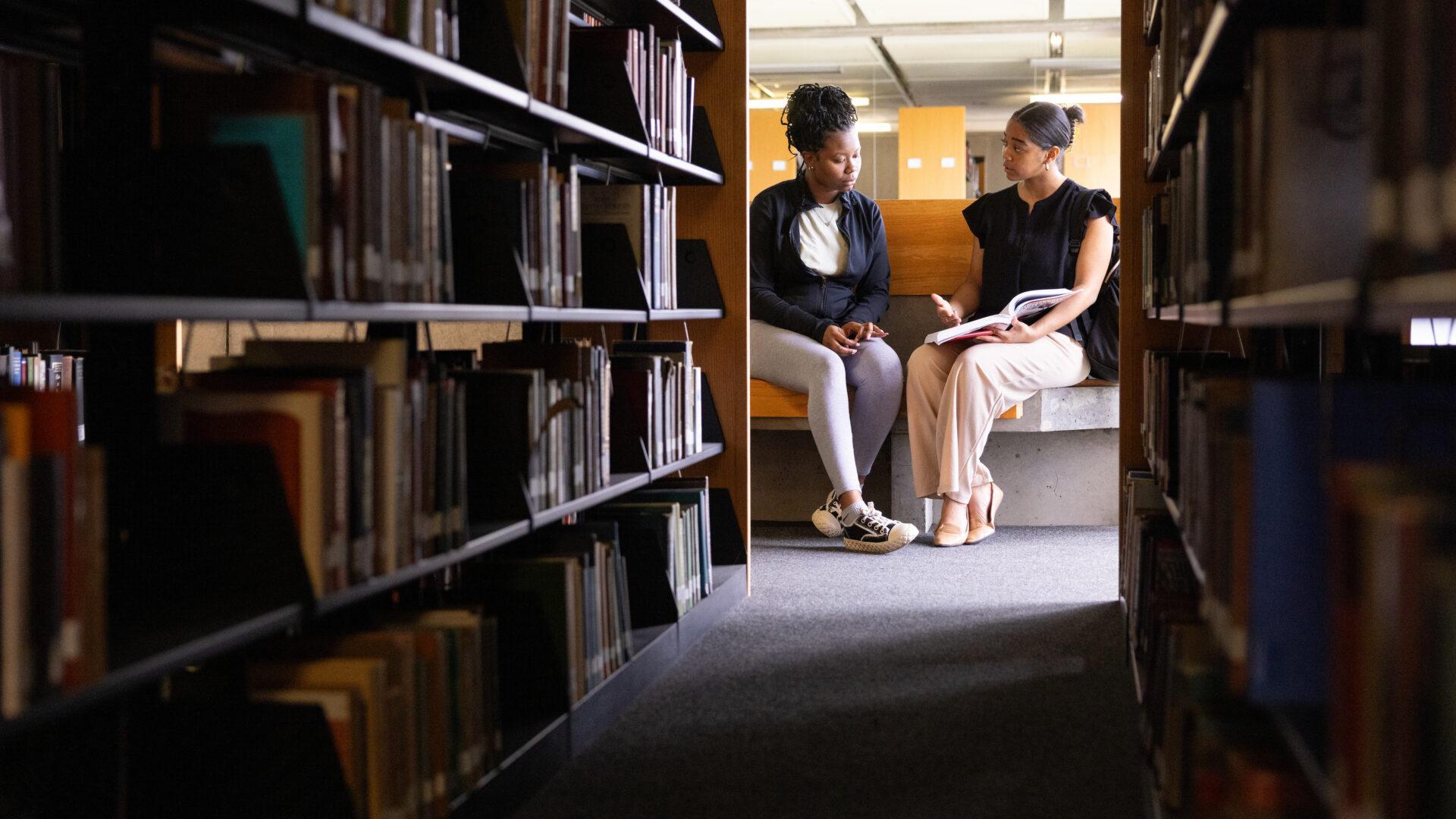 Two Black female students looking at a book together at the end of a row at a college library.