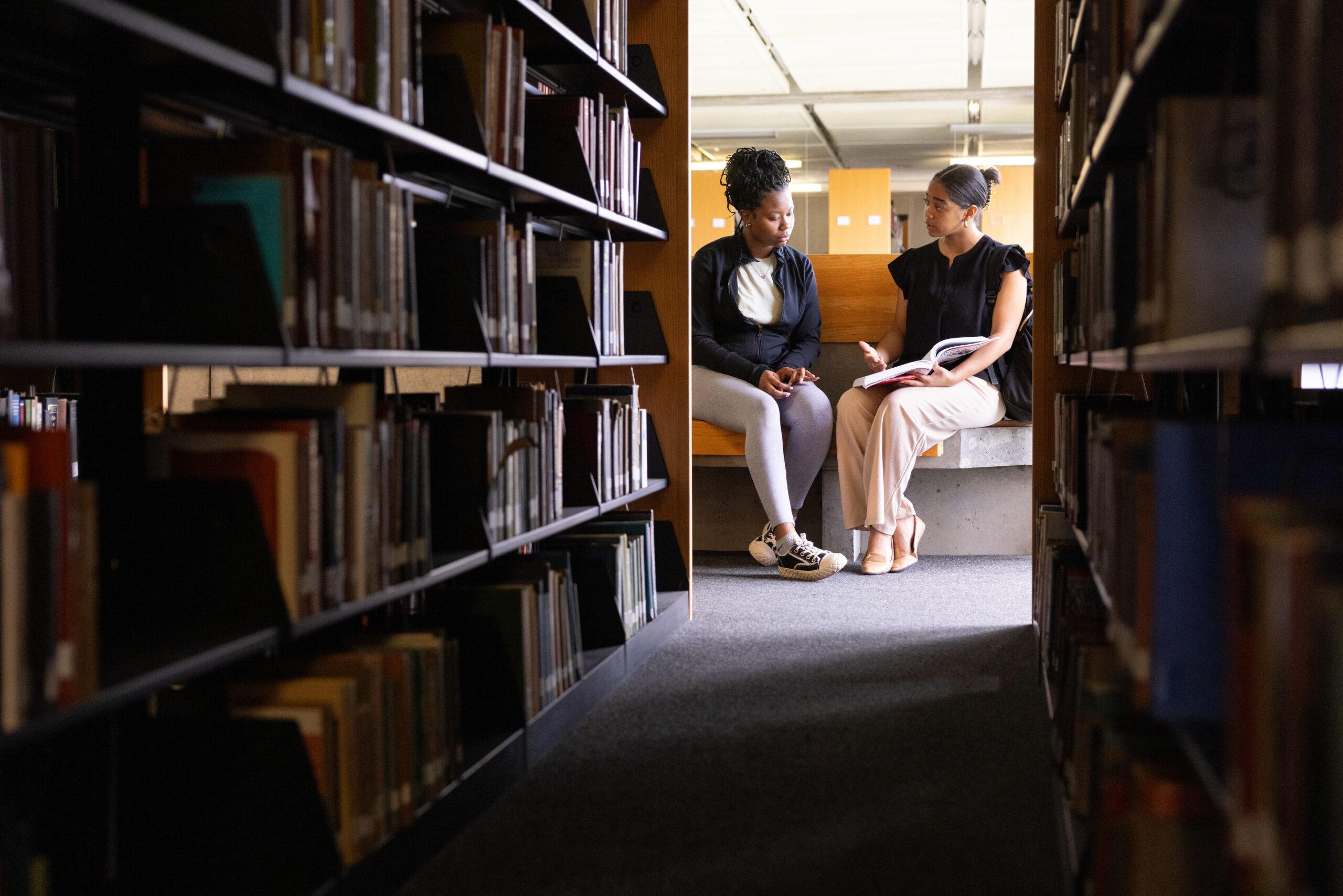 Two Black female students looking at a book together at the end of a row at a college library.