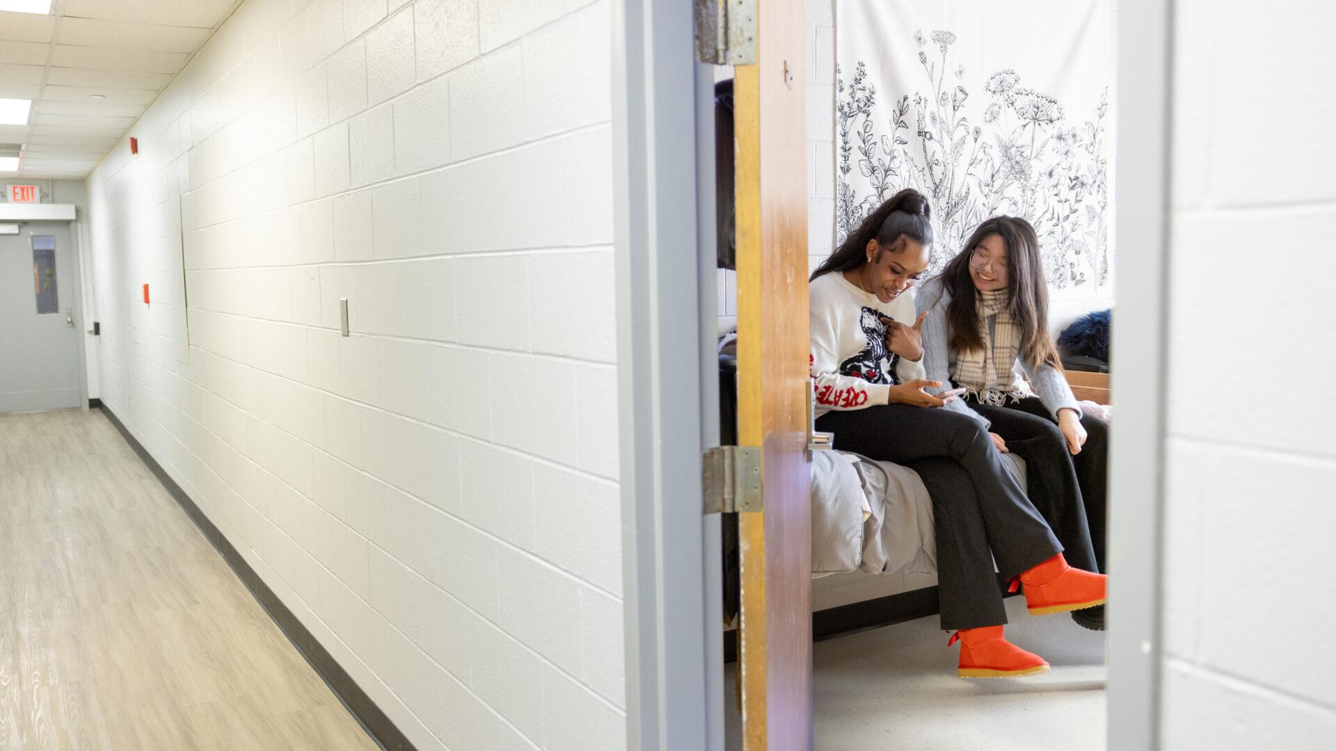Two female students looking at a phone in a dorm room on a college campus.