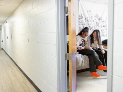 Two female students looking at a phone in a dorm room on a college campus.