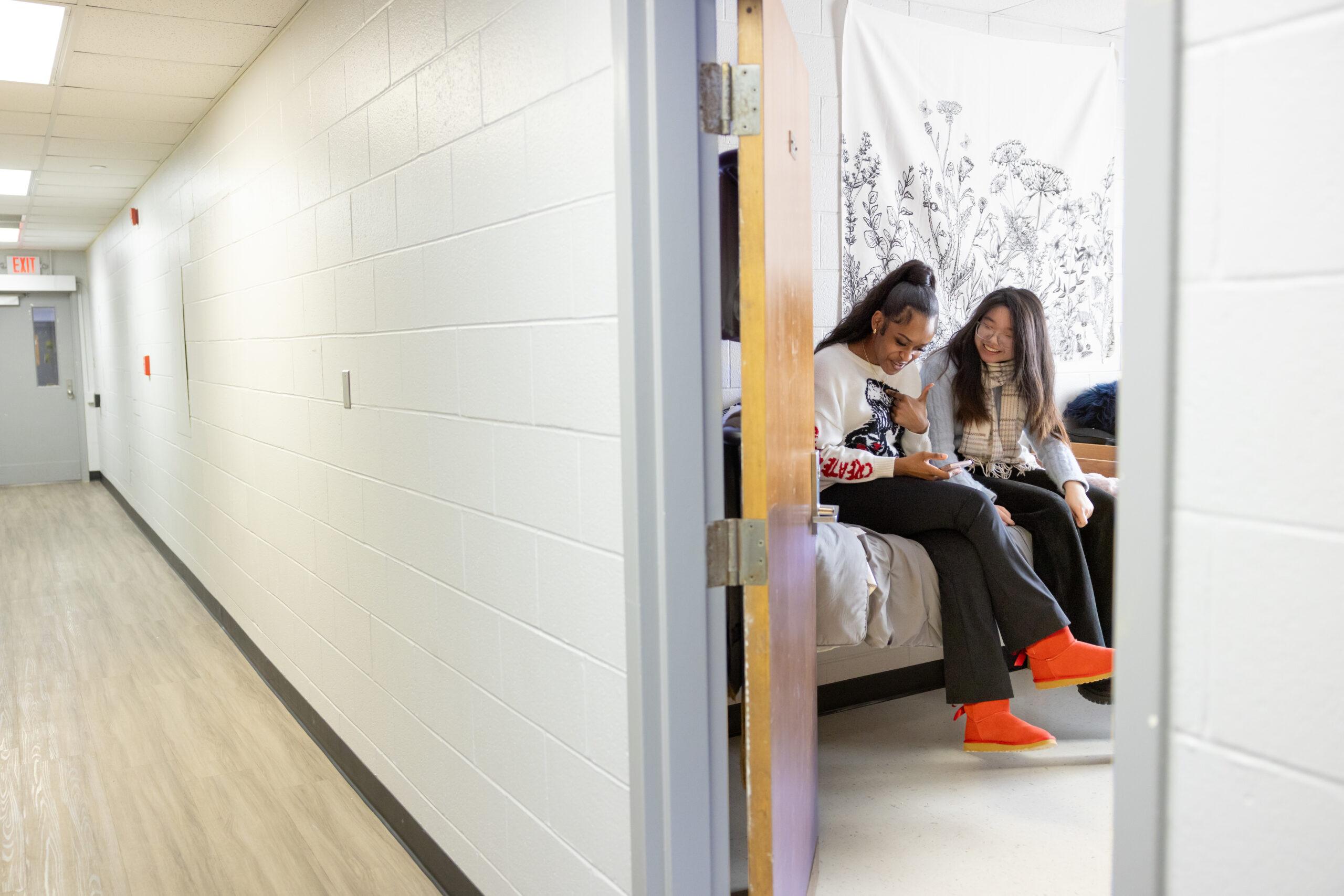 Two female students looking at a phone in a dorm room on a college campus.
