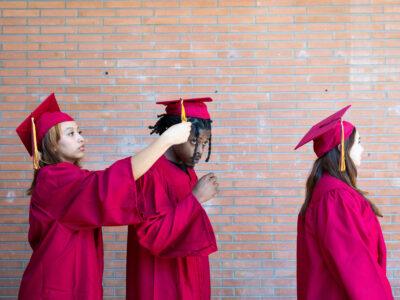 Two Hispanic female students in graduation cap and gown adjusting the mortarboard of a Black male student in preparation for a graduation ceremony on a college campus.