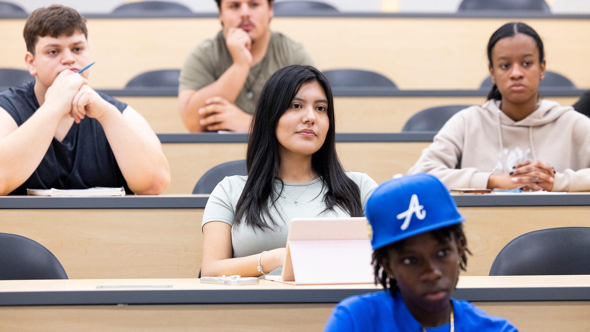 Hispanic female student listening in a large classroom with fellow students on a college campus.