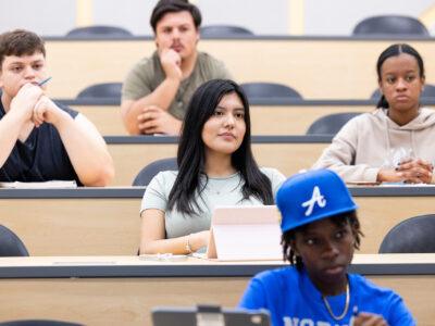 Hispanic female student listening in a large classroom with fellow students on a college campus.